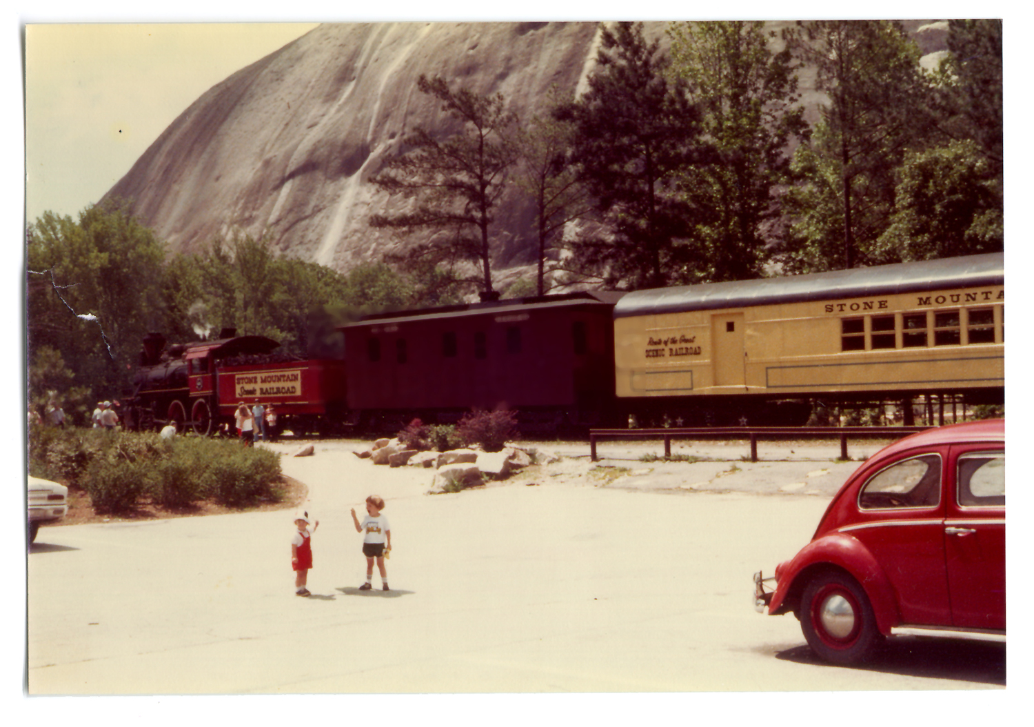 A photograph from the 1970s with two children standing in front of a passing train with part of a red Volkswagen bug in the foreground and mountains in the distance. 